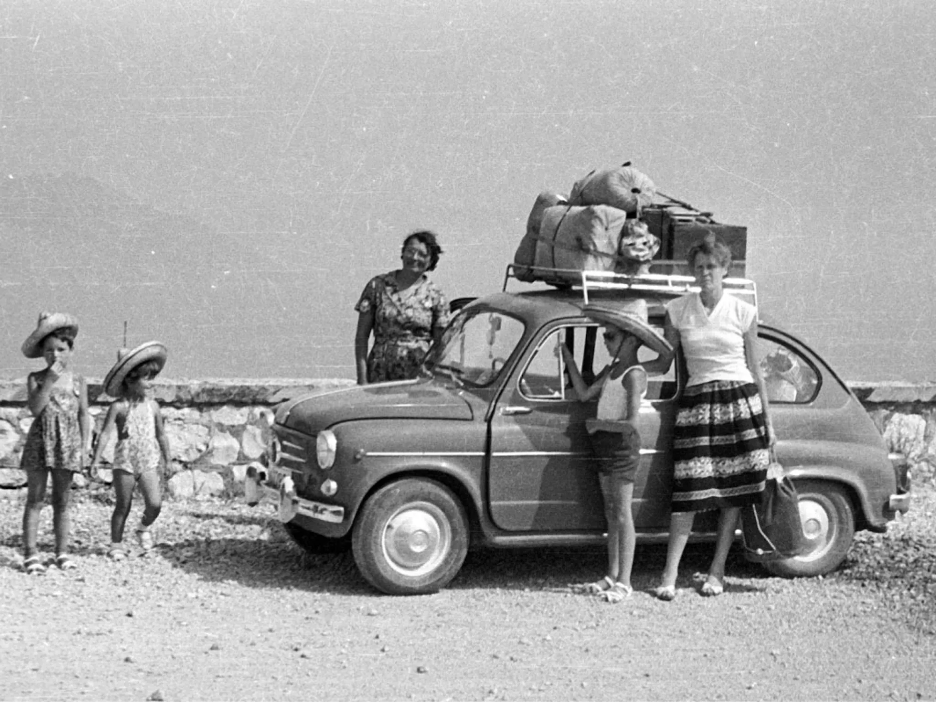 Stara črno-bela fotografija družine pred avtom, na strehi je privezana prtljaga